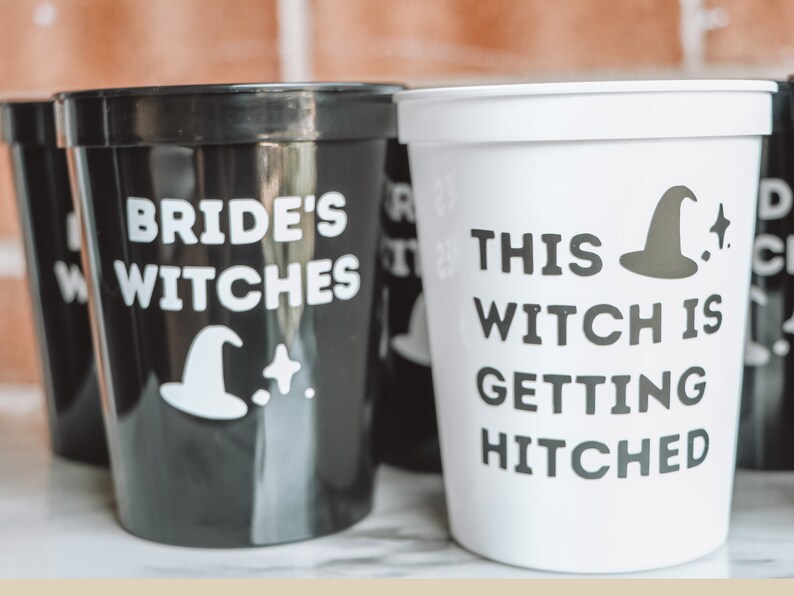 May include: Black and white plastic cups with text and witch hat graphics. One cup reads "Bride's Witches" and another says "This Witch Is Getting Hitched". The cups are likely for a bachelorette party or bridal shower.