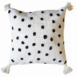 Op de afbeelding: Een wit kussen met zwarte stippen en kwastjes aan elke hoek.