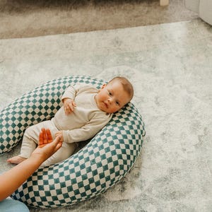 以下が含まれることがあります： ベージュの服を着た赤ちゃんが、緑と白のチェック柄の授乳枕の上にいます。枕はU字型で、サポートを提供します。赤ちゃんは明るい色のラグの上に横たわっています。