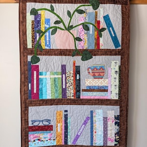 May include: A quilted wall hanging depicting a bookshelf with books and a potted plant. The bookshelf is brown, with three shelves filled with colorful fabric books. A pink pot with green leaves and vines is in the center. A pair of glasses rests on the bottom shelf.