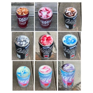 Może przedstawiać: Kolekcja dziewięciu świec w puszkach po napojach, w tym Dr. Pepper, Oreo Coca-Cola Zero Sugar i Coca-Cola Y3000. Każda świeca ma unikalny, kolorowy wz&oacute;r wosku na wierzchu z widocznym knotem umieszczonym centralnie wewnątrz wosku.
