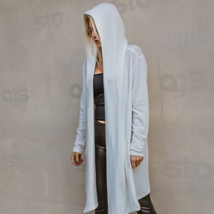 May include: A white hooded cardigan with long sleeves and a long, flowing design. The cardigan is worn over a black tank top and black leggings. The person is wearing black heeled boots.