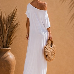 May include: A floor-length white maxi dress with short sleeves and an off-the-shoulder neckline. The dress features a loose, flowing silhouette and is paired with a round, textured straw handbag with fringe detailing and tan platform sandals.