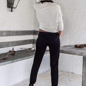 Puede incluir: Una mujer con una blusa blanca de manga larga con cuello negro y pantalones negros. Lleva botines negros de tac&oacute;n alto. El fondo incluye una pared blanca, un banco de piedra y una mesa de madera.