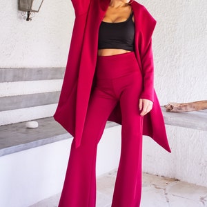 Op de afbeelding: Een vrouw draagt een bordeauxrood broekpak met een zwart topje en een zonnebril. De broek is wijd en het jasje heeft een lang, vloeiend ontwerp.