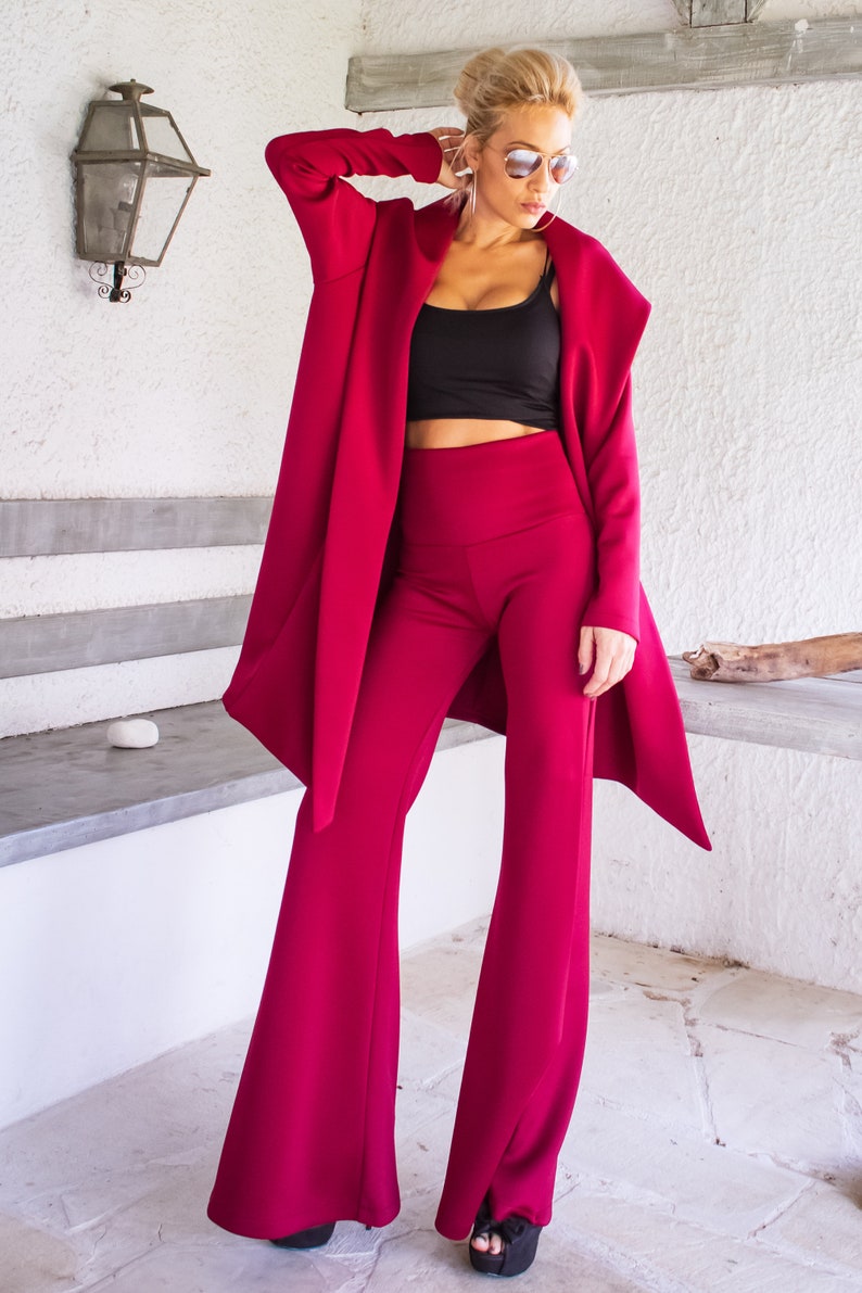 Op de afbeelding: Een vrouw draagt een bordeauxrood broekpak met een zwart topje en een zonnebril. De broek is wijd en het jasje heeft een lang, vloeiend ontwerp.