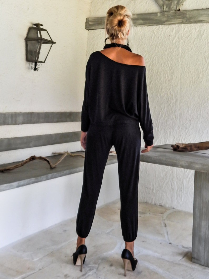 May include: A woman wearing a black off-the-shoulder jumpsuit with a relaxed fit. The jumpsuit has long sleeves and a slightly cropped leg. She is wearing black high heels.