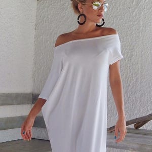 May include: A solid white off-the-shoulder dress with short sleeves and a loose, flowing silhouette. The garment features a wide neckline that rests across the shoulders, creating a relaxed drape that extends down to the mid-calf area.