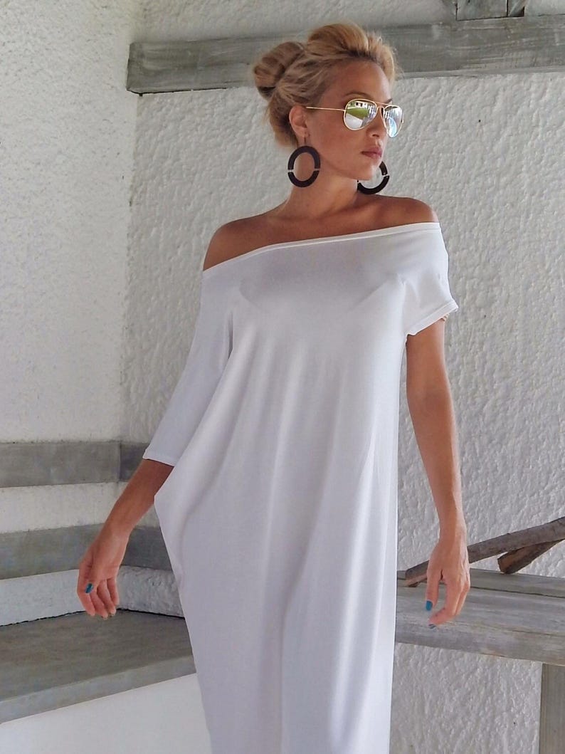 May include: A solid white off-the-shoulder dress with short sleeves and a loose, flowing silhouette. The garment features a wide neckline that rests across the shoulders, creating a relaxed drape that extends down to the mid-calf area.
