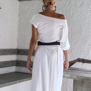 May include: A white off-the-shoulder maxi dress featuring a loose, draped bodice with short sleeves and a long, flowing skirt. The garment is cinched at the waist with a wide, solid black belt that features a rectangular gold-toned metal buckle.