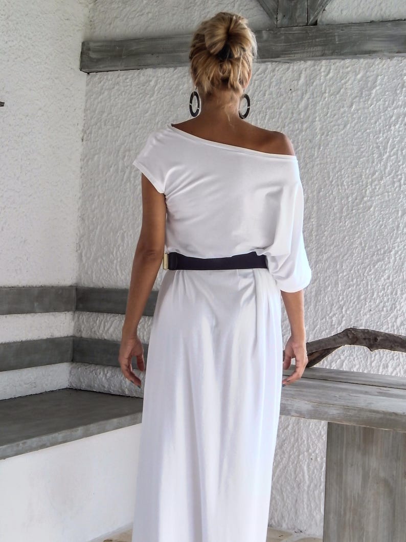 May include: A white off-the-shoulder maxi dress featuring a loose, draped bodice with short sleeves and a long, flowing skirt. The garment is cinched at the waist with a wide, solid black belt that features a rectangular gold-toned metal buckle.