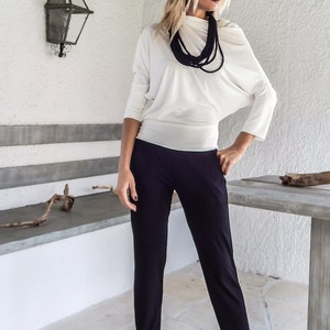 Puede incluir: Una mujer con una blusa blanca de manga larga con cuello drapeado y pantalones negros. Un collar negro y gafas de sol completan el atuendo. La mujer est&aacute; de pie afuera, cerca de una pared blanca y una mesa de madera.