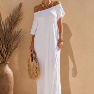 May include: A floor-length white maxi dress featuring an off-the-shoulder neckline and short sleeves. The garment is paired with a round straw handbag with wooden handles, gold-toned layered necklaces, gold earrings, and platform sandals.