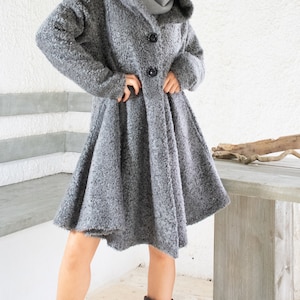 Könnte beinhalten: Ein grauer Mantel mit Kapuze und ausgestelltem Rock, mit großen schwarzen Knöpfen. Der Mantel wird mit einem grauen Schal und dunkelgrauen Stiefeln kombiniert. Ein stilvolles Outfit für kaltes Wetter.