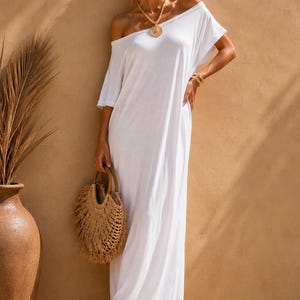 May include: A floor-length white off-the-shoulder dress with short sleeves, paired with a round woven straw handbag featuring fringe detailing, gold-toned circular drop earrings, a beaded necklace with a circular pendant, and platform sandals.