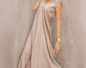 May include: A beige, sleeveless dress with a V-neck. The dress has a flowing, A-line silhouette and falls to the ankles. The model is wearing sunglasses and high-heeled sandals. The background is a neutral, textured wall.