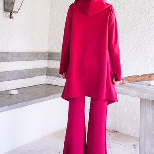 Op de afbeelding: Een magenta hooded jas en bijpassende wijde broek. De jas heeft een lang, vloeiend ontwerp en de broek heeft een uitlopende pasvorm.