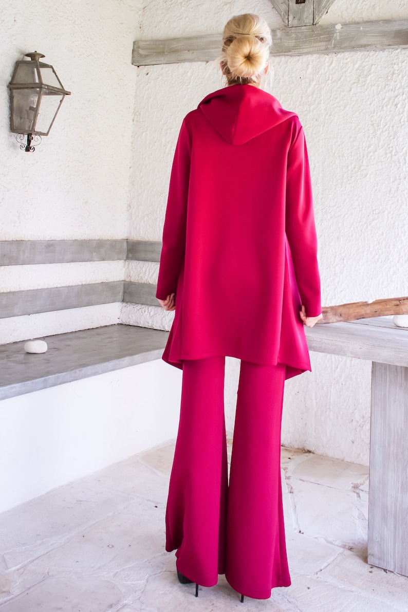 Op de afbeelding: Een magenta hooded jas en bijpassende wijde broek. De jas heeft een lang, vloeiend ontwerp en de broek heeft een uitlopende pasvorm.
