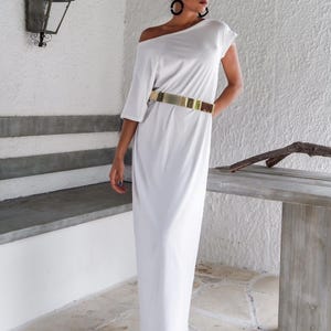 May include: A floor-length white dress with an off-the-shoulder neckline and short sleeves. The dress is cinched at the waist with a wide, metallic gold-toned belt. The garment features a smooth, solid fabric construction and a straight, flowing silhouette.