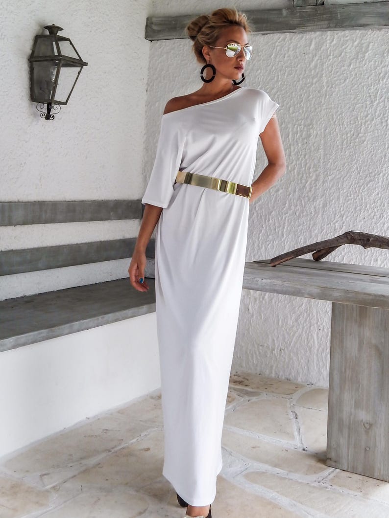 May include: A floor-length white dress with an off-the-shoulder neckline and short sleeves. The dress is cinched at the waist with a wide, metallic gold-toned belt. The garment features a smooth, solid fabric construction and a straight, flowing silhouette.