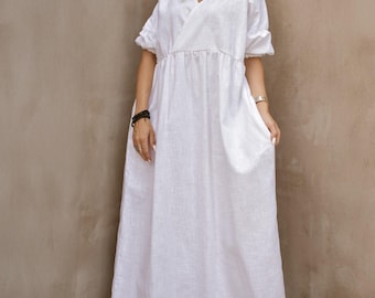 May include: A white linen maxi dress with a V-neckline and short sleeves. The dress features a gathered waist and a flowing skirt. The model is wearing sunglasses and platform sandals. The dress is a summer or resort wear.