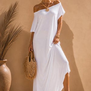 May include: A solid white maxi dress featuring a loose, flowing silhouette with an off-the-shoulder neckline and short sleeves. The garment is paired with a round woven straw handbag with wooden handles and tan platform sandals with ankle straps.