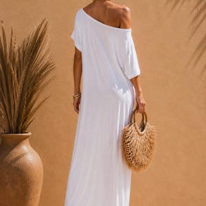 May include: A floor-length white maxi dress with short sleeves and an off-the-shoulder neckline. The dress features a loose, flowing silhouette and is paired with a round, textured straw handbag with fringe detailing and tan platform sandals.