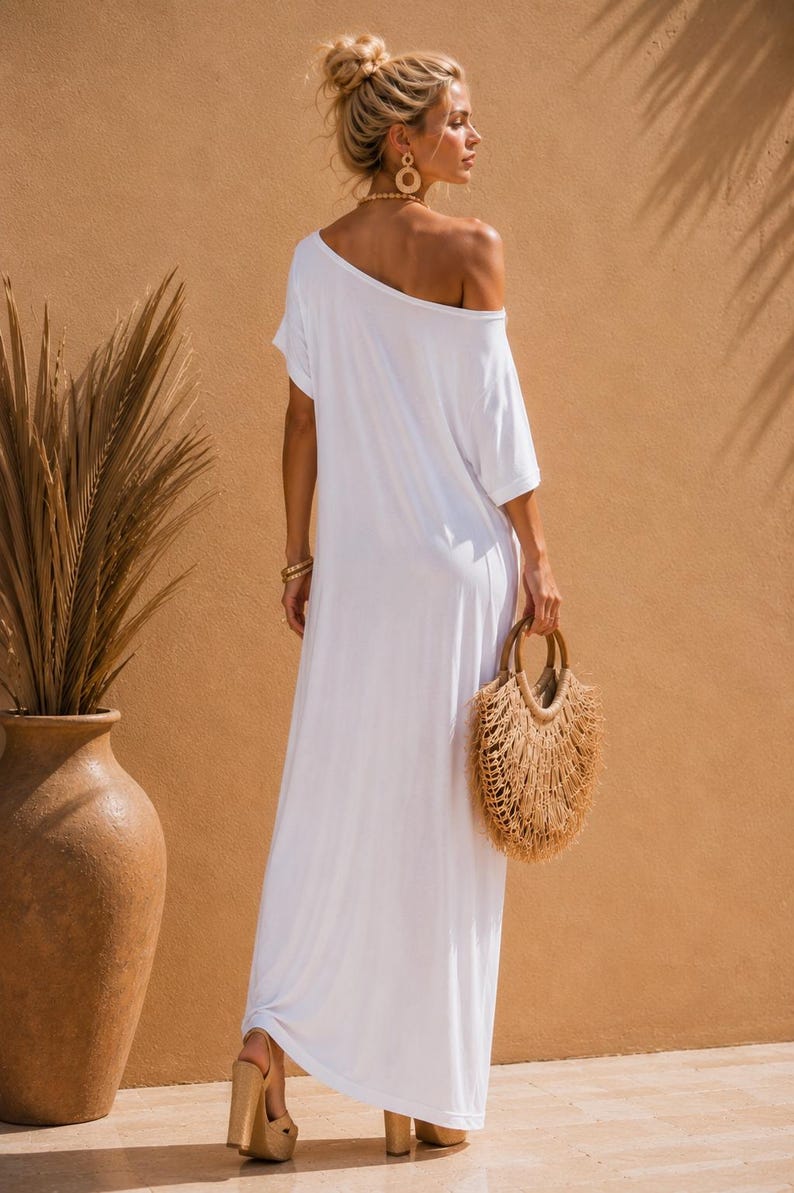 May include: A floor-length white maxi dress with short sleeves and an off-the-shoulder neckline. The dress features a loose, flowing silhouette and is paired with a round, textured straw handbag with fringe detailing and tan platform sandals.