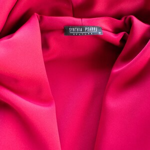 Op de afbeelding: Een close-up van een felroze kledingstuk met capuchon met een zwart label waarop "SYNTHIA PSARRU COUTURE" en "M" staat.