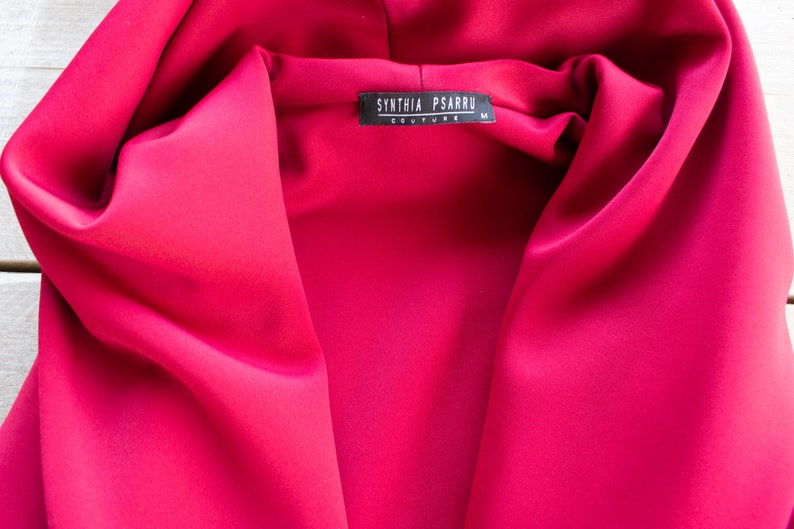 Op de afbeelding: Een close-up van een felroze kledingstuk met capuchon met een zwart label waarop "SYNTHIA PSARRU COUTURE" en "M" staat.