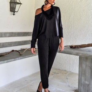 May include: A woman wearing a black one-shoulder jumpsuit with a black scarf necklace and sunglasses. She is standing in front of a white wall with a wooden beam above her head.