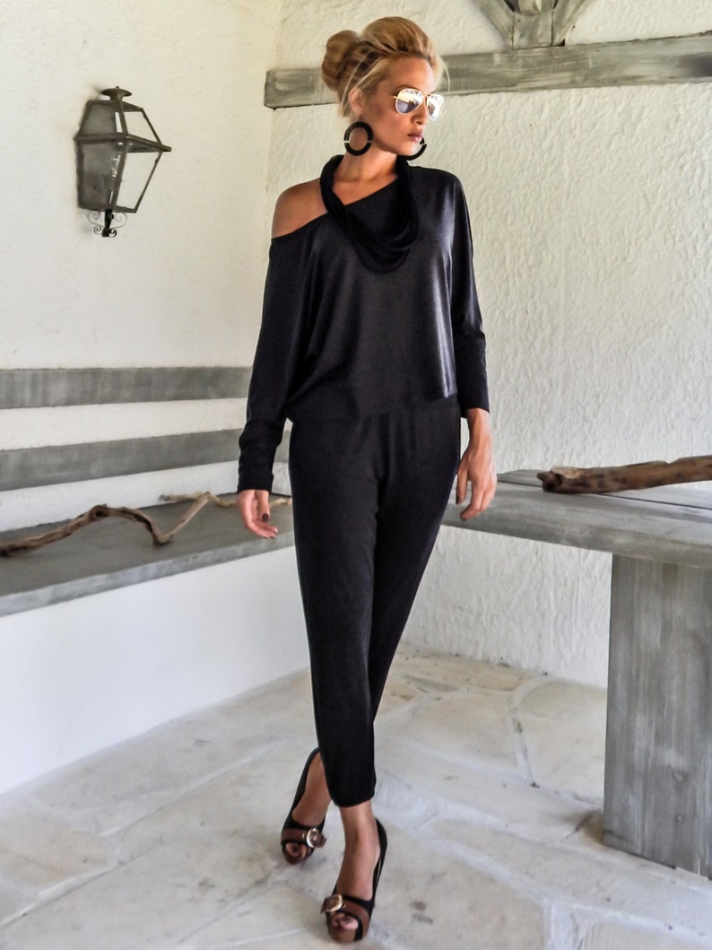 May include: A woman wearing a black one-shoulder jumpsuit with a black scarf necklace and sunglasses. She is standing in front of a white wall with a wooden beam above her head.