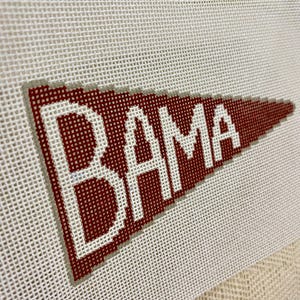 May include: A needlepoint pennant in crimson red with the word "BAMA" in white block letters. The pennant has a gray border and is set against a white canvas background.