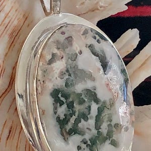 May include: Oval pendant with a silver-coloured metal setting. The pendant features a large, polished stone with a pattern of white, green, and brown inclusions. The bail is a simple loop. The pendant is displayed on a seashell.
