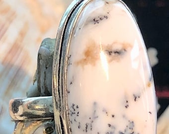 May include: A silver ring with an oval-shaped white agate stone with black and brown inclusions. The stone is set in a silver band with a smooth, polished finish.