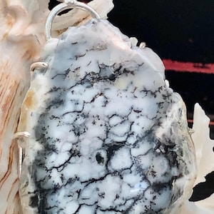 May include: A silver pendant featuring a white and black dendritic agate stone. The stone is set in a silver bezel and has a natural, organic shape.