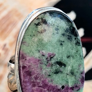 Peut inclure: Une bague en argent avec une grande pierre ovale verte et violette. La pierre présente un motif marbré de vert, de violet et de noir.