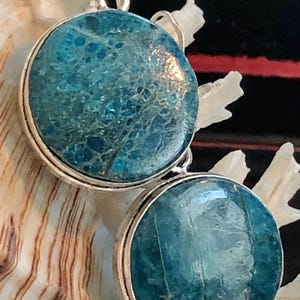 May include: Round, turquoise-colored earrings with a silver-colored metal border. The earrings have a marbled appearance and are suspended from a silver-colored hook. The background is a seashell.