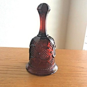 Könnte beinhalten: Eine dunkelrote Glasglocke mit einer strukturierten Oberfläche und einem dekorativen Design. Die Glocke hat eine abgerundete Basis und einen hohen, schlanken Griff.