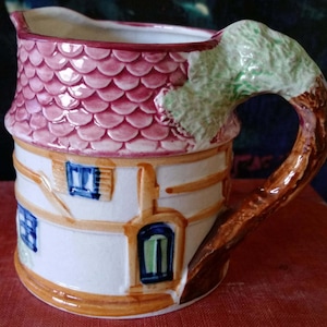 Puede incluir: Una jarra de cerámica blanca con forma de casa con un techo rosa y un asa de árbol verde. La casa tiene dos ventanas y una puerta.