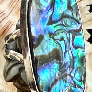 May include: A ring featuring a large, elongated, oval-shaped abalone shell inlay with iridescent blue, green, and purple tones. The shell is set within a polished silver-toned metal bezel and attached to a split, textured silver-toned metal band.