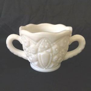 Puede incluir: Azucarero blanco de vidrio de leche con dos asas y un borde festoneado. El azucarero tiene un patrón en relieve.