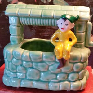 May include: A green ceramic planter shaped like a well with a small elf figurine sitting on the edge. The elf is wearing a yellow shirt and a green hat.