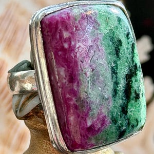 May include: A large, rectangular ring with a silver band. The focal point is a vibrant gemstone, showcasing a blend of deep magenta and emerald green hues. The stone is set within a silver bezel.