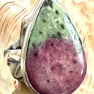 Peut inclure: Une bague en forme de goutte d'eau ornée d'une pierre de zoïsite polie présentant une séparation de couleurs. La partie supérieure est vert clair moucheté avec des inclusions sombres, tandis que la base est d'un magenta-pourpre profond.