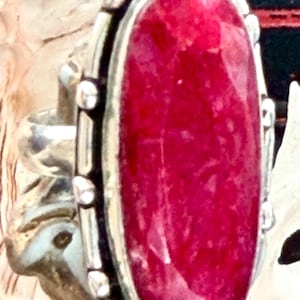 May include: A ring featuring a large, oval-shaped, deep pinkish-red faceted gemstone set in a silver bezel. The metal setting is detailed with small, evenly spaced silver beads along the perimeter of the stone, secured by a simple silver band.