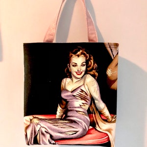 May include: A rectangular fabric tote bag featuring a graphic print of a person with long brown hair wearing a purple dress and white gloves, seated on a red cushion against a black background. The bag has two light pink glittery handles.