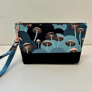 May include: A small, rectangular pouch with a black bottom and a teal and black mushroom pattern. The pouch has a rose gold zipper and a matching wrist strap. The mushroom design features light pink and white details.