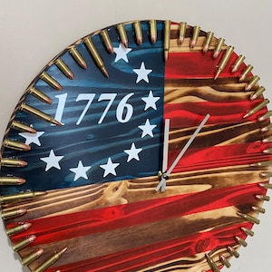 May include: A round wooden clock with a red, white, and blue American flag design. The clock is surrounded by a ring of brass bullet casings. The clock face features the number 1776 in white lettering.