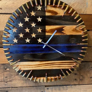 May include: A wooden clock with a thin blue line and an American flag design. The clock is surrounded by bullet casings.